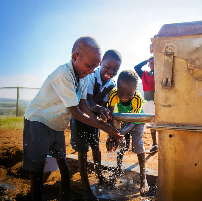 three smiling African children drink water from a borehole
