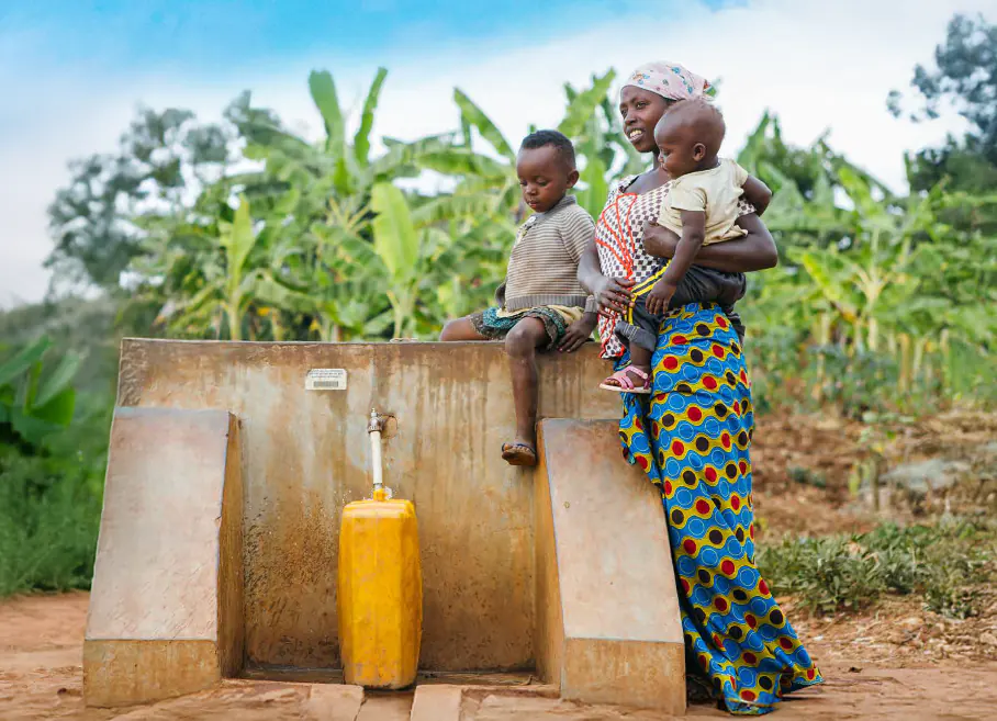 A young African woman holds her baby next to a water point while collecting water, her son sat beside her